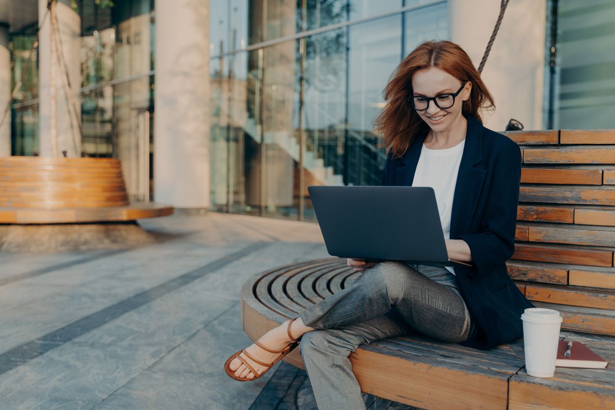 kobieta siedzi przed biurowcem z laptopem na kolanach