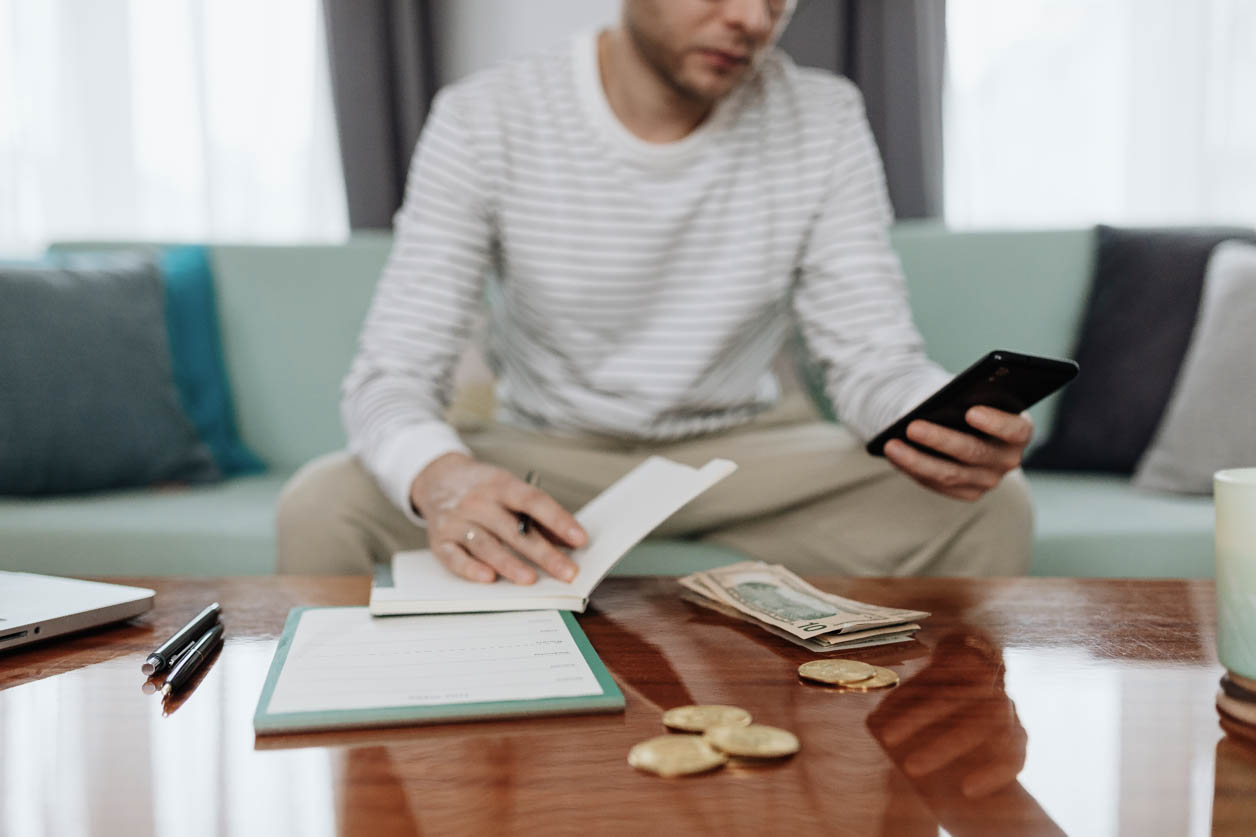 Młody mężczyzna oblicza wysokość rachunków na telefonie i w notatniku, pieniądze leżą na stole